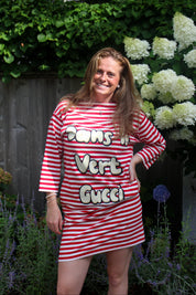 A woman posing outdoors in a red and white striped t-shirt dress with 'Dans un Vert Gucci' printed across the front, set against a background of green foliage and white flowers.