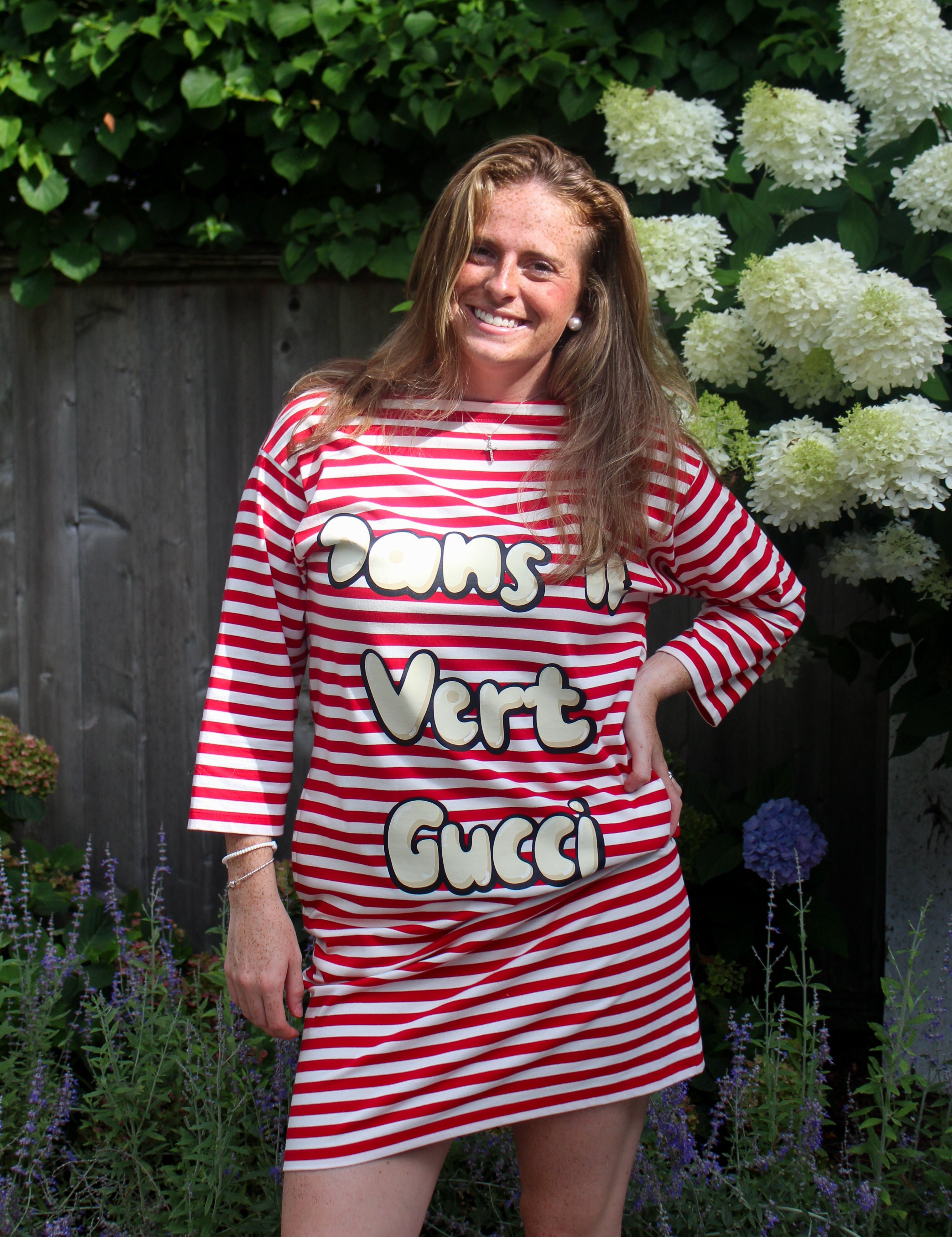 A woman posing outdoors in a red and white striped t-shirt dress with 'Dans un Vert Gucci' printed across the front, set against a background of green foliage and white flowers.
