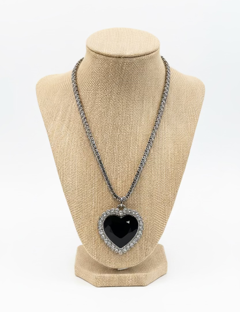 A heart-shaped pendant necklace with a black center and a silver rhinestone border, displayed on a beige fabric jewelry bust. The necklace is set against a busy retail background with a green and white patterned tablecloth.