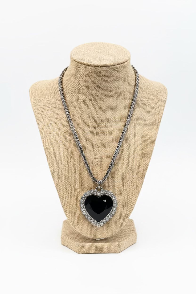 A heart-shaped pendant necklace with a black center and a silver rhinestone border, displayed on a beige fabric jewelry bust. The necklace is set against a busy retail background with a green and white patterned tablecloth.